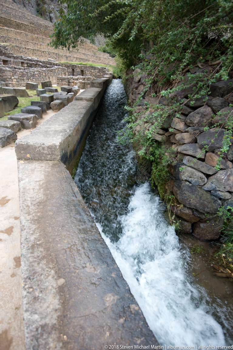 Fast-moving water in a narrow stone-lined channel bordered by masonry and vegetation at Ollantaytambo.