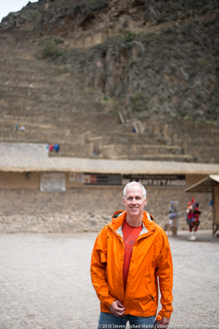 Visitor in an orange jacket standing in an open plaza with Ollantaytambo ruins and terraces behind.