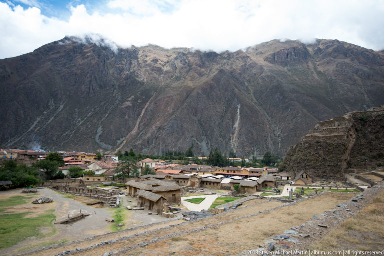 Wide view over lower ruins and lawns toward Ollantaytambo town and steep mountains, with stone terrace remains at right.