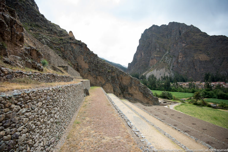Long level path beside a stone retaining wall, with narrow descending terraces, green fields, and mountains beyond.