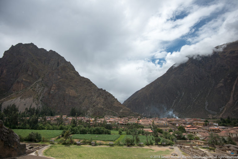 Wide view of Ollantaytambo town and green fields beneath steep mountain slopes and a cloud-filled sky.