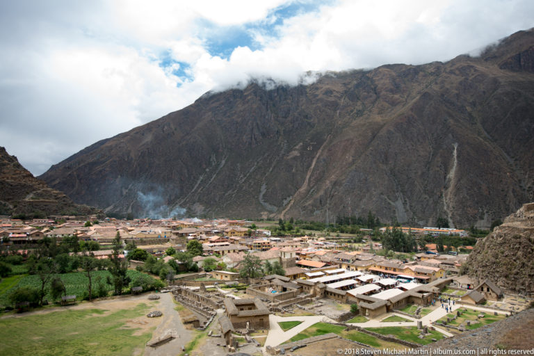 Elevated overview of lower stone foundations and open courts with Ollantaytambo town and a steep mountain slope behind.