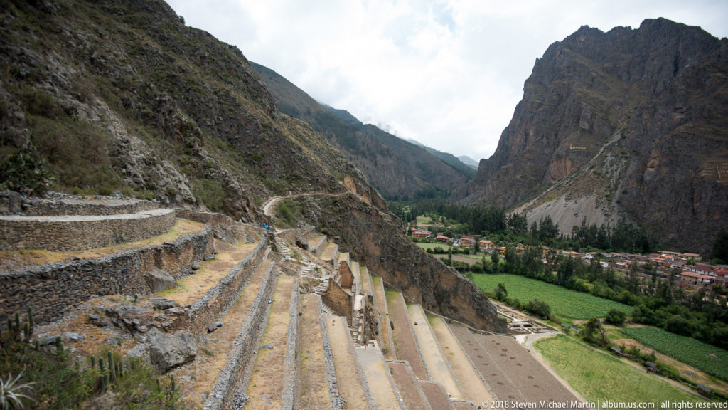 Long parallel stone terraces descend a steep slope toward green fields and the town of Ollantaytambo below.