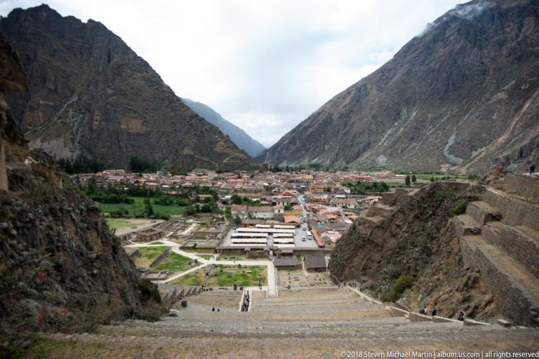High view down over Ollantaytambo ruins, paths, town roofs, green fields, and steep mountains enclosing the valley.