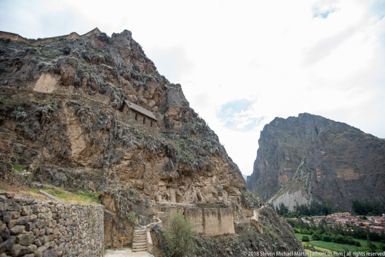 Roofed stone storehouse high on a rocky hillside, seen above lower walls, paths, and vegetation near Ollantaytambo.