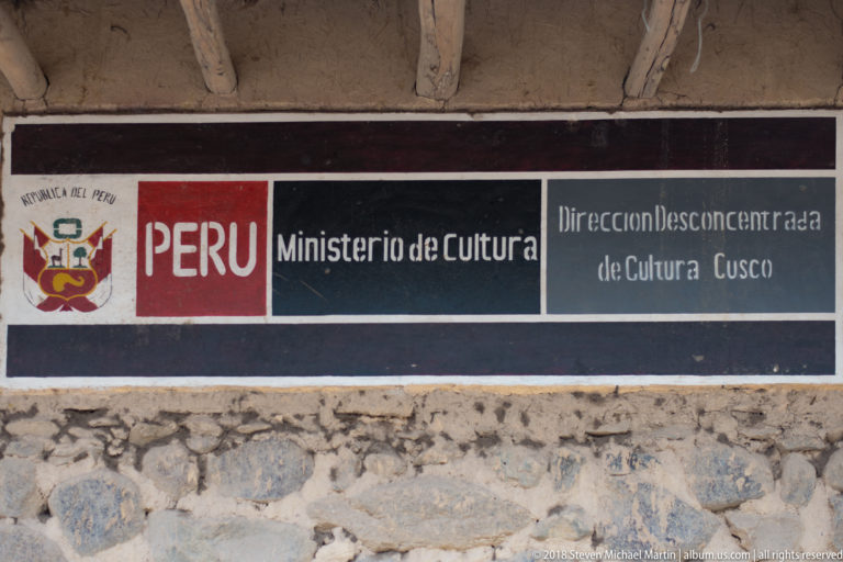 Official Peru Ministry of Culture sign mounted on a stone-and-earth wall at Ollantaytambo.