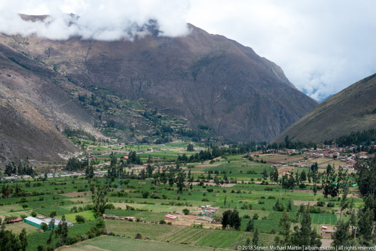 Green agricultural fields and scattered settlement spread across a valley floor beneath steep cloud-covered Andean mountains.