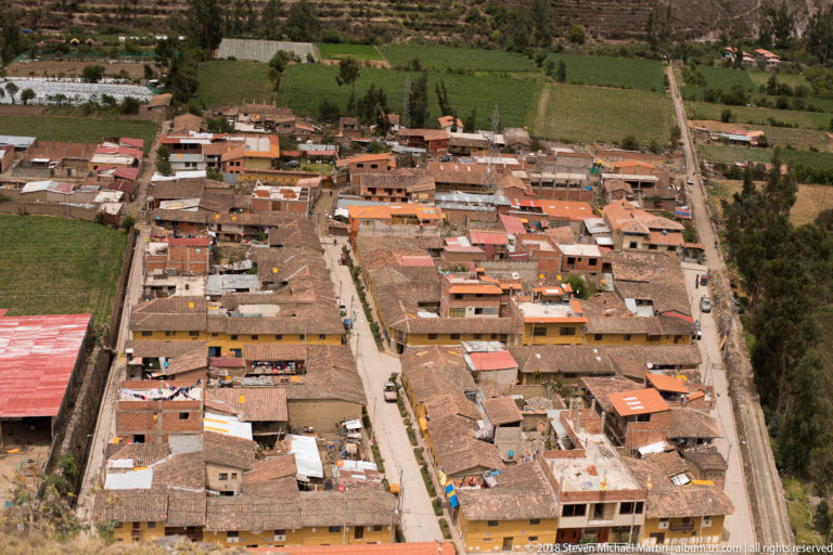 Elevated view over Ollantaytambo town, showing compact roofed blocks, straight streets, trees, and cultivated fields on the valley floor.