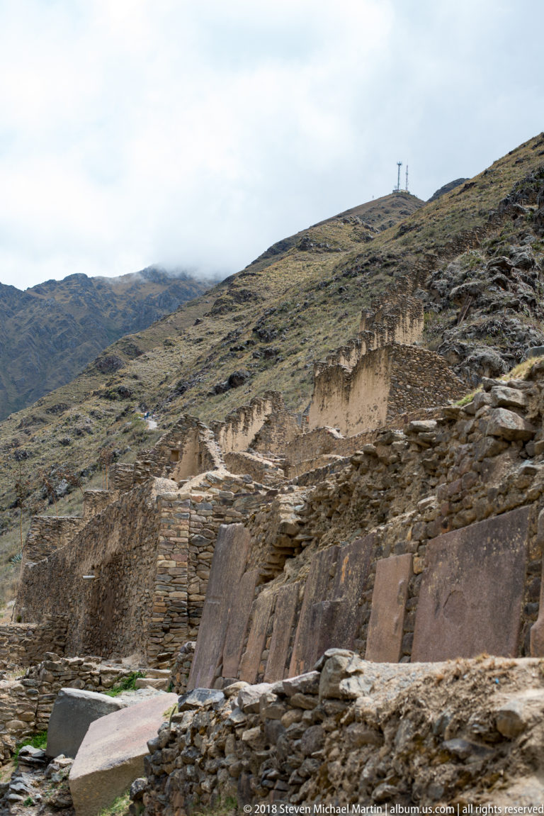 Wide view of ruined stone walls and terraces on a mountainside, with several large dressed slabs among the foreground masonry.
