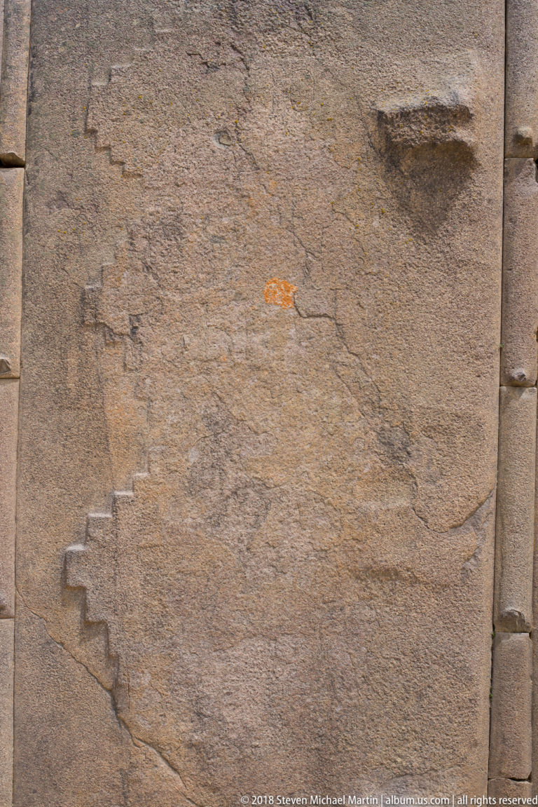 Close-up of a weathered dressed stone face with shallow rectangular recesses, a crack, and a small patch of orange lichen.