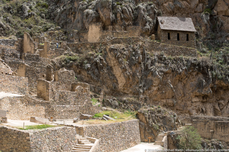 Stone terraces and stairs fill the lower slope beneath a small roofed masonry storehouse built high into the rocky cliff.