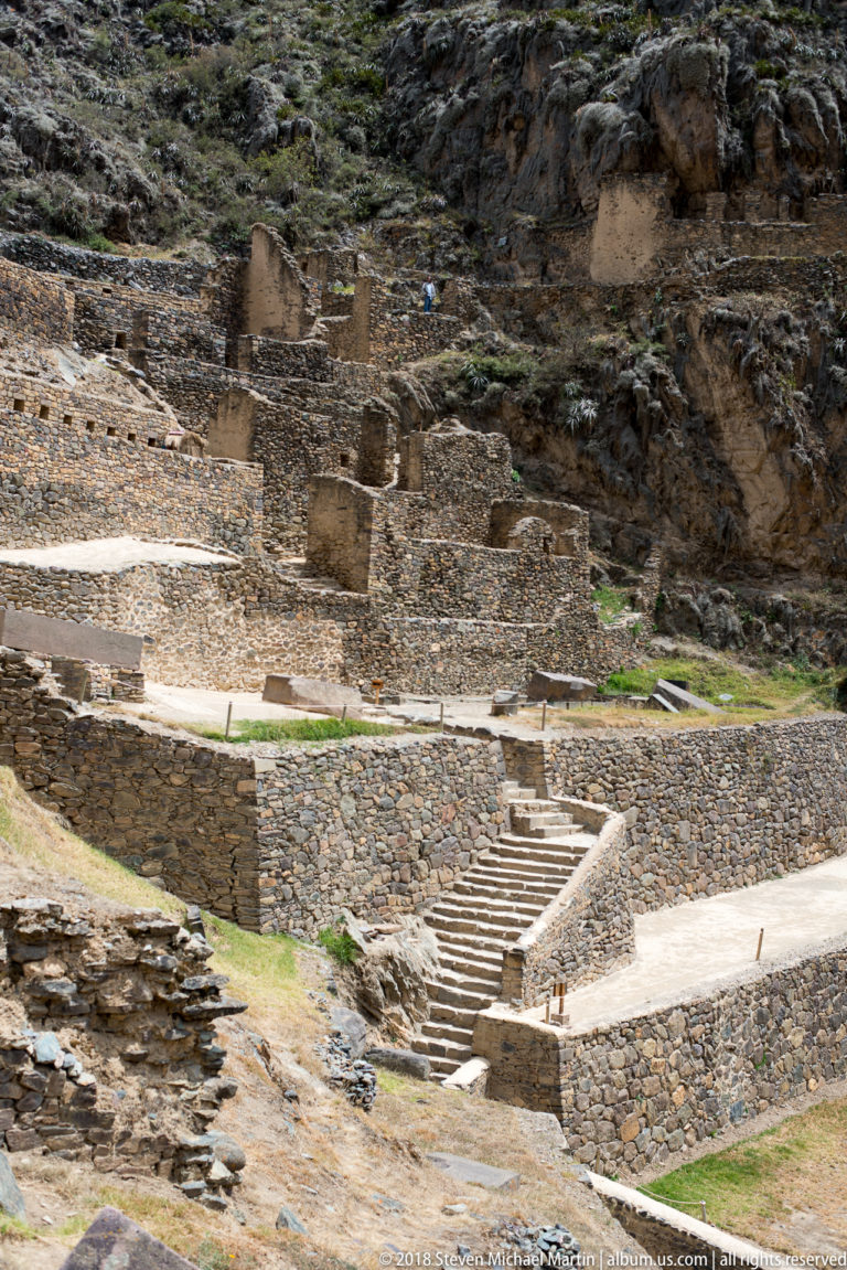 Broad stone stairway rises between multiple stone retaining walls and level terraces on a rocky archaeological slope.