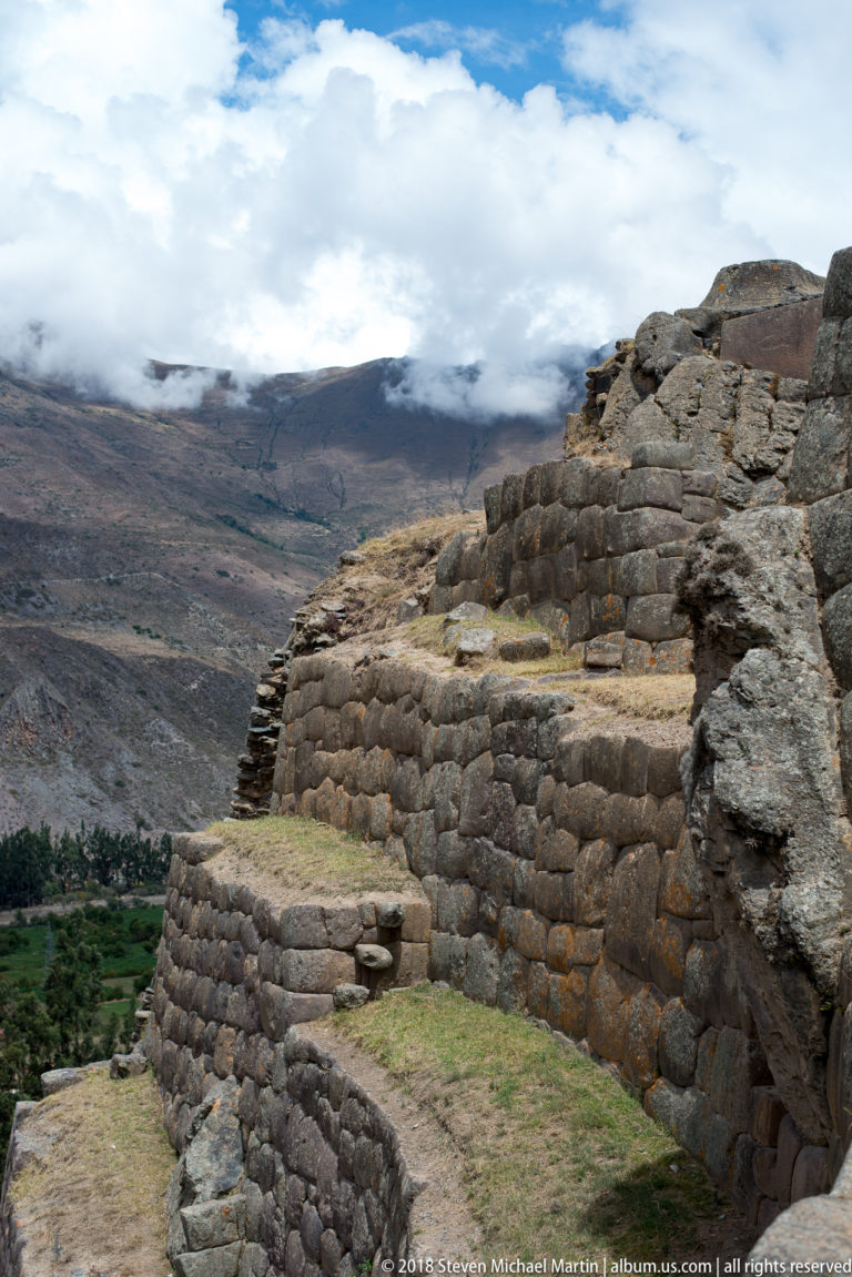 Stepped stone retaining walls descend along a high slope above fields and broad cloud-covered mountains.