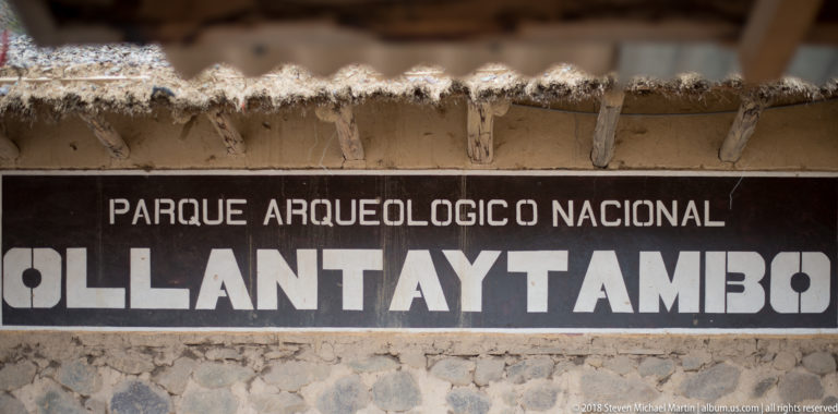 Large painted sign reading “Parque Arqueológico Nacional Ollantaytambo” beneath a rustic roof.