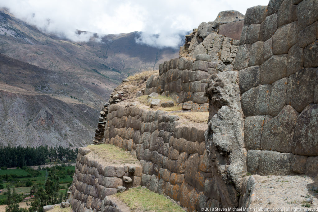Large stone terrace walls on a high slope overlooking green fields and the mountain valley below Ollantaytambo.