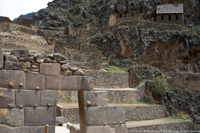 Dressed-stone doorway in front of rising terraces, with a small roofed stone storehouse visible high on the rocky cliff.