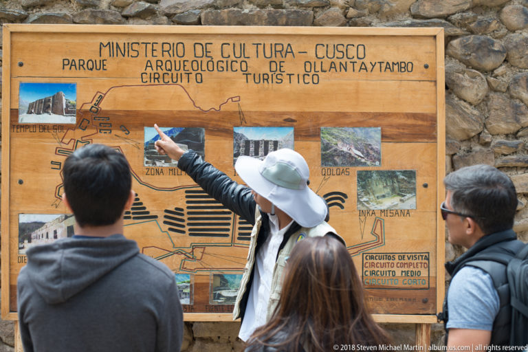 Guide in a sun hat points to a large Ollantaytambo archaeological circuit map while several visitors stand in front of it.