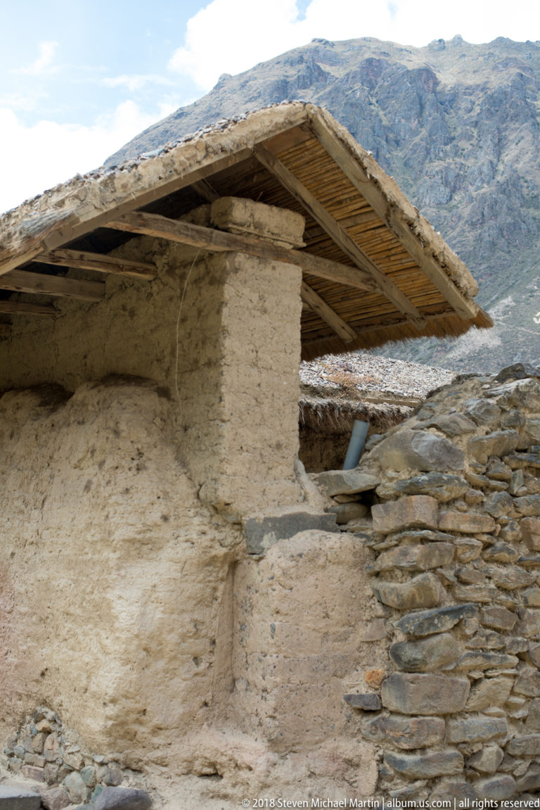 Close view of a thick earthen wall under a rustic pitched roof, beside rough stone masonry with mountains in the background.
