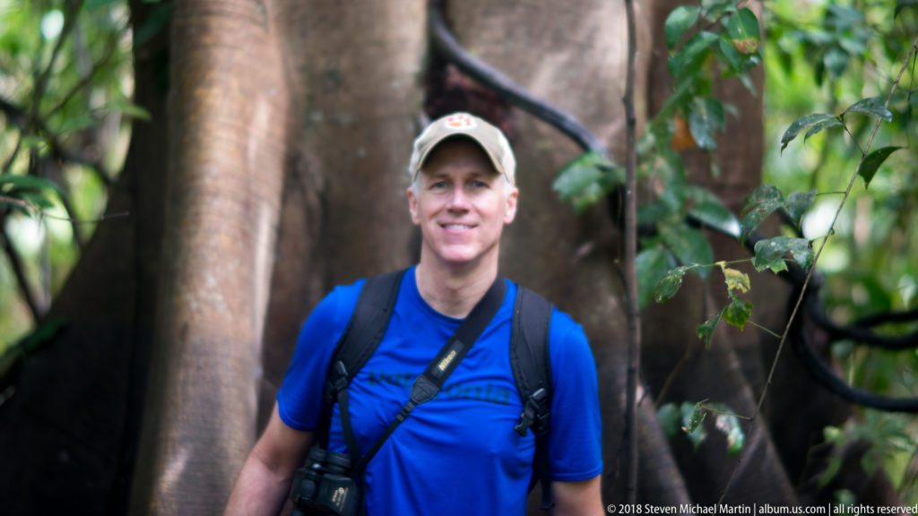 Upper-body portrait of a visitor with binoculars and backpack in front of smooth giant tree buttresses.
