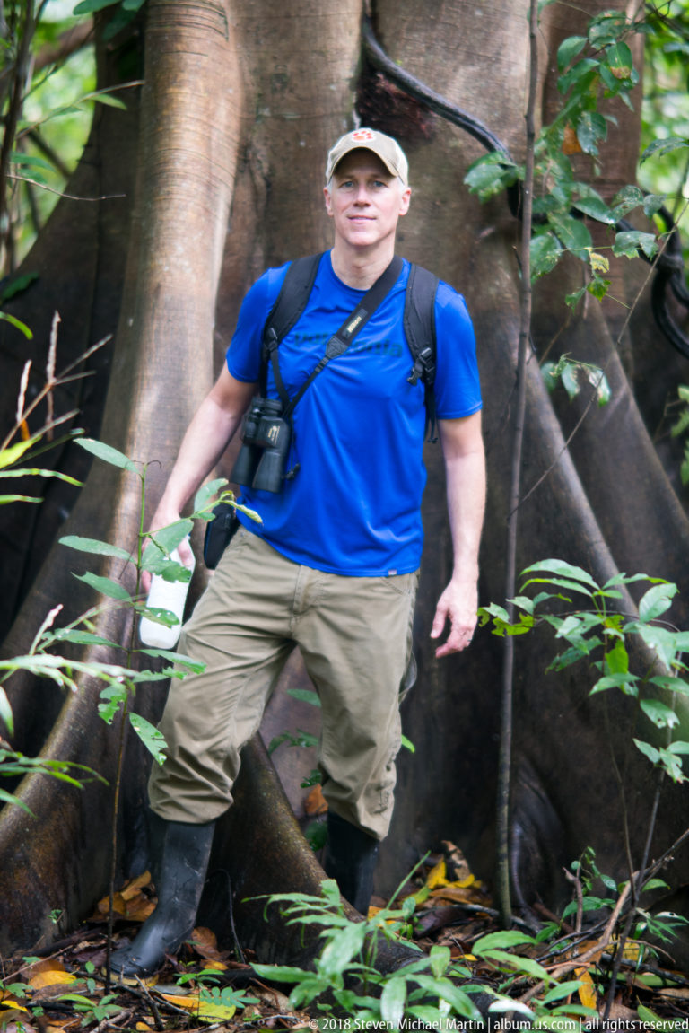 Full-length visitor in field gear standing beside broad tree buttresses amid leaf litter and understory plants.