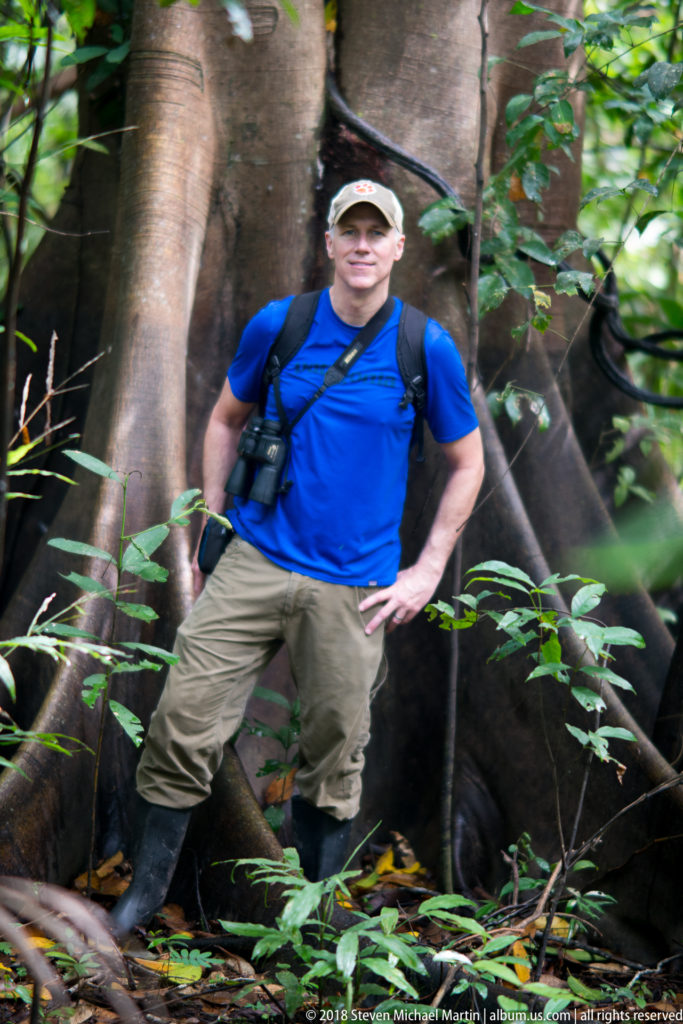 Centered portrait of a visitor standing before towering buttress roots with binoculars and backpack.