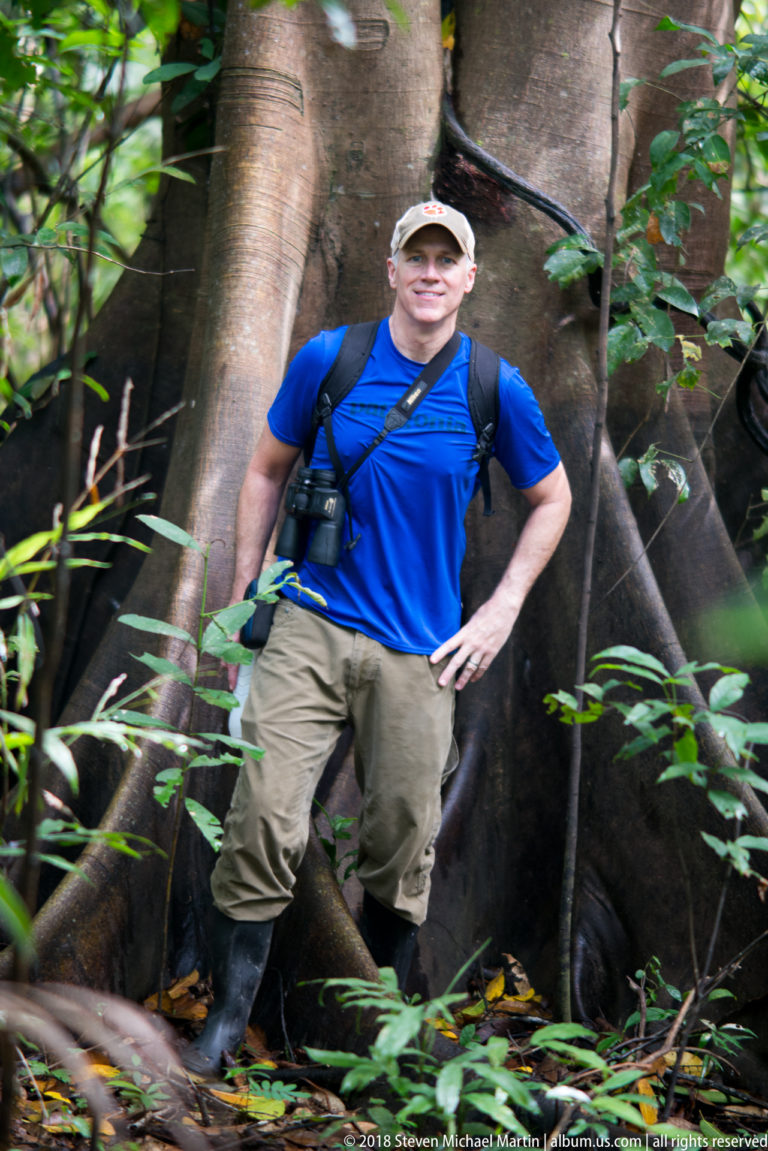 Visitor with backpack and binoculars standing beside massive buttress roots in dense Amazon rainforest.
