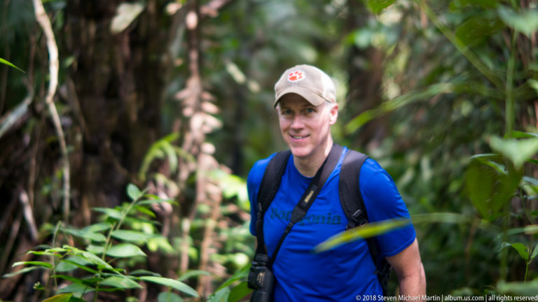 Man in blue shirt and cap standing among palms, vines and broad leaves in dense rainforest.