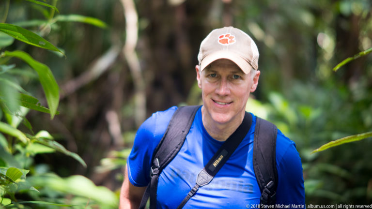 Man in blue shirt, cap and backpack standing among dense green rainforest vegetation.
