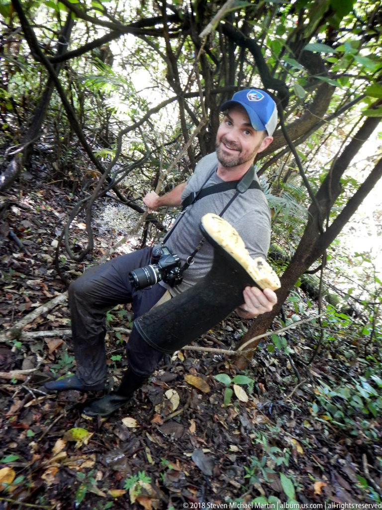 Man with camera crouching in rainforest understory while holding a large pale shelf fungus toward the camera.