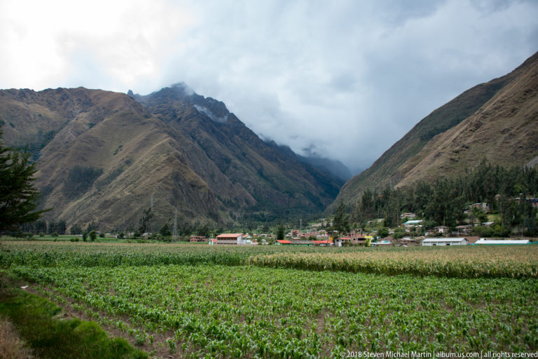 Broad green field leading to a small settlement below steep cloud-covered Andean mountains.
