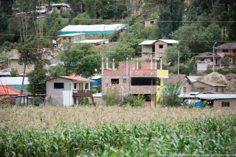 Rural houses, utility poles and unfinished brick construction behind a tall cornfield in an Andean valley.