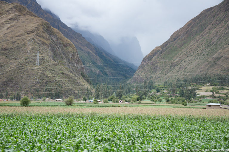 Green crop field on a flat valley floor between steep brown Andean mountains fading into cloud.