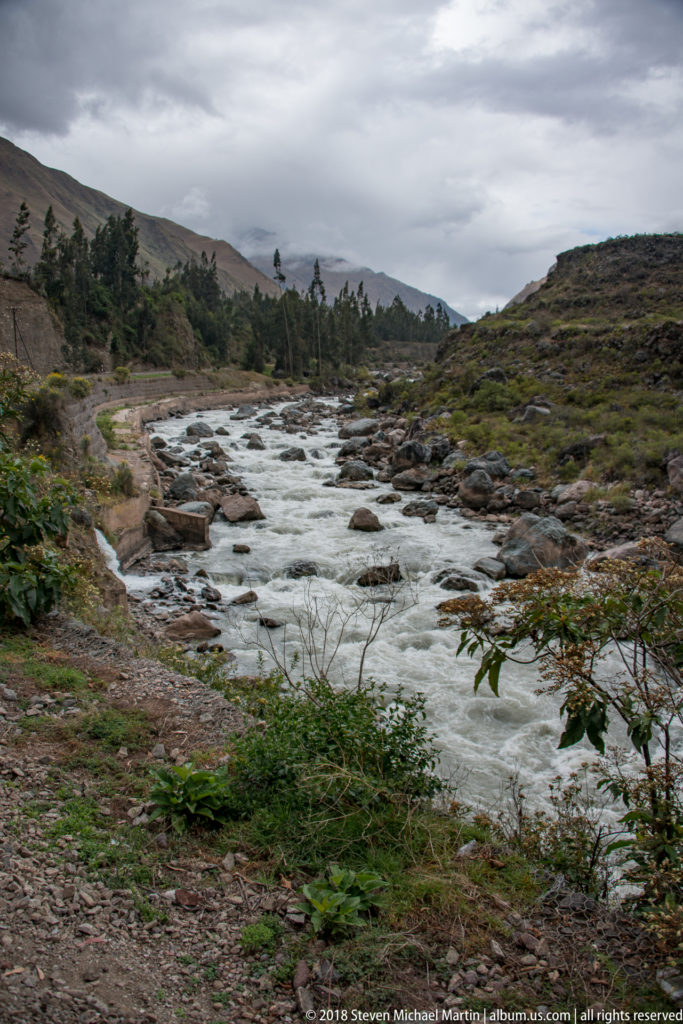Shallow rocky river with many exposed stones flowing through an open Andean valley under clouds.