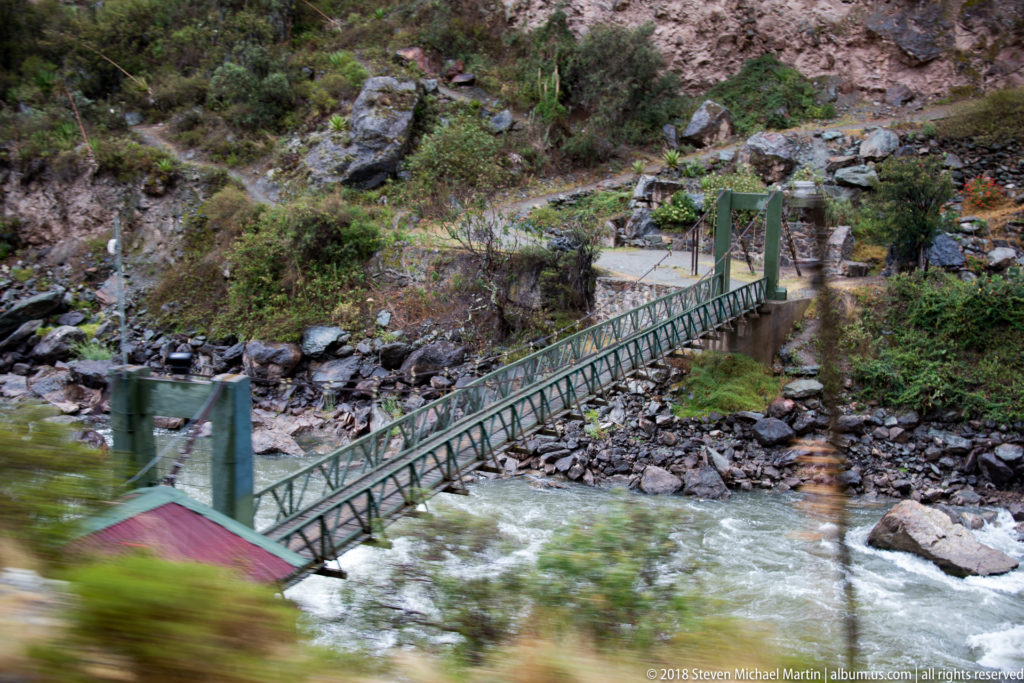 Diagonal metal suspension footbridge over rushing water, seen with motion blur from a passing train.