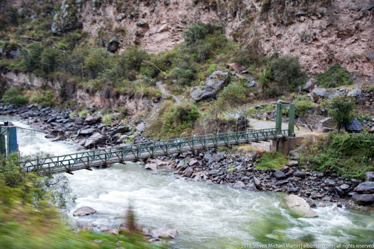 Narrow metal suspension footbridge crossing a turbulent rocky river below a steep hillside.