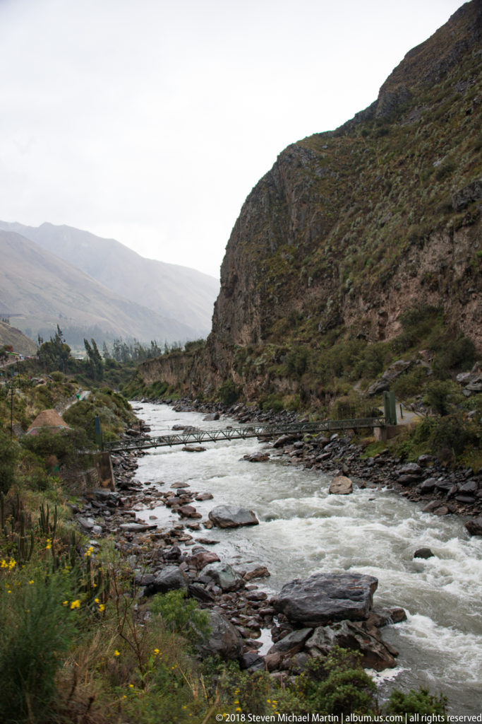 Fast-flowing rocky river between sparsely vegetated Andean slopes under an overcast sky.