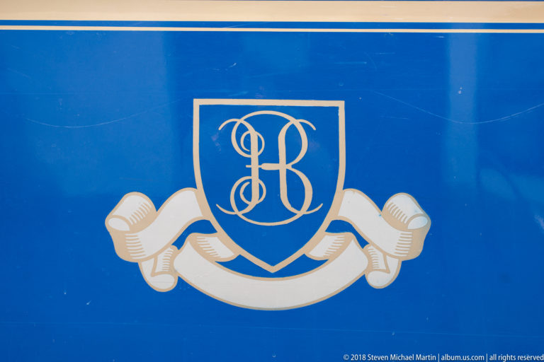 Gold monogram crest and ribbon motif painted on the deep blue side of a luxury passenger railcar.