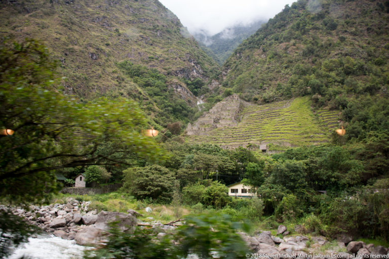 Stepped green agricultural terraces above a rocky river in a steep Andean valley, seen from a train.