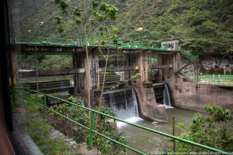 Concrete spillway with rushing water, green railings and service walkways in a steep green valley.