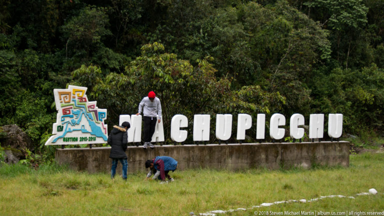 Several visitors pose around and on top of a large MACHUPICCHU sign beside forest.