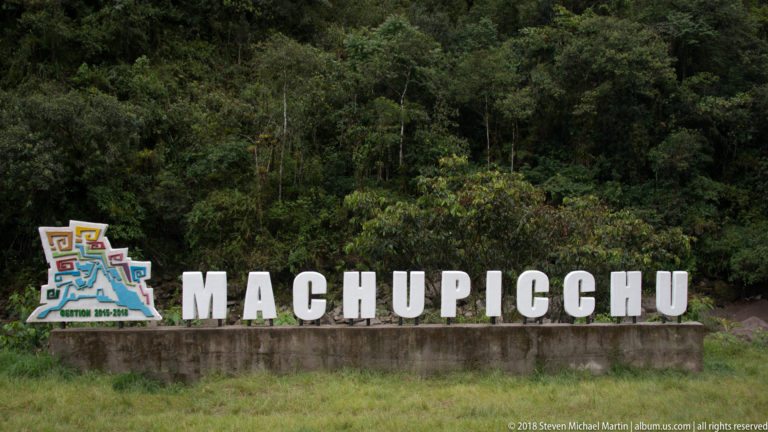Large white MACHUPICCHU sign with a colorful anniversary emblem in front of dense forest.