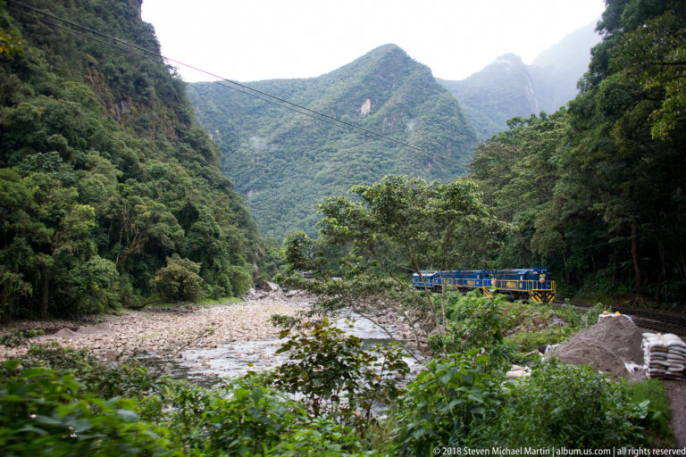 Blue-and-yellow passenger train beside a rocky river in a densely forested Andean gorge.