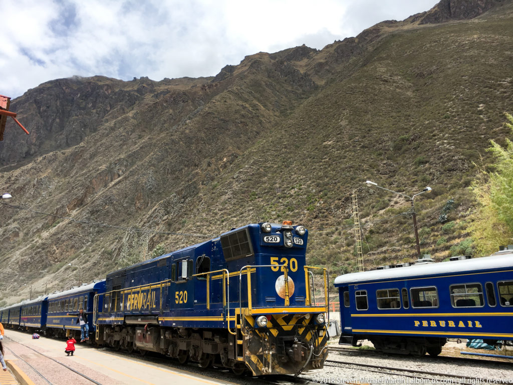 Blue-and-yellow PeruRail locomotive 520 with passenger coaches beneath steep Andean slopes.