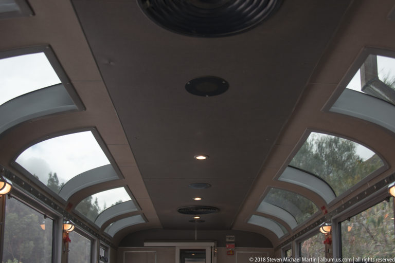 Interior of a sightseeing train car with curved panoramic roof windows, side glazing and ceiling lights.