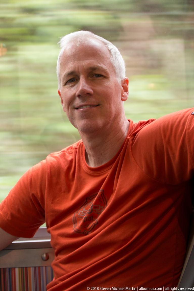 Smiling seated passenger in an orange shirt beside a train window with blurred scenery outside.