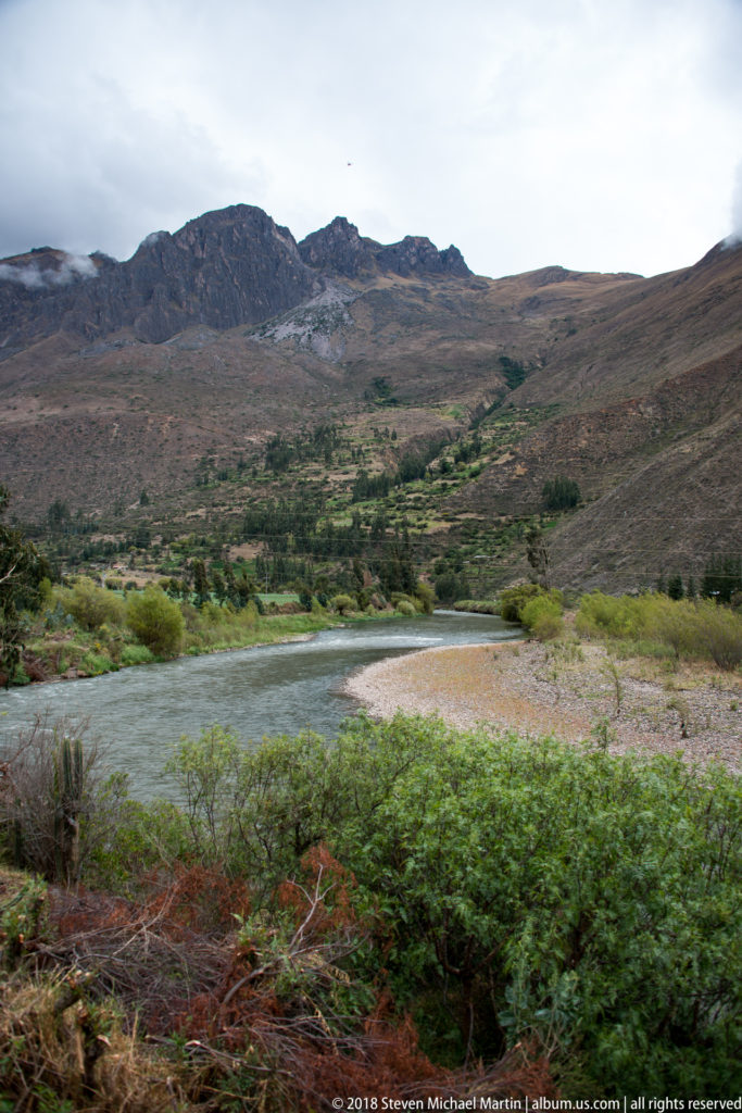 Wide river bend and gravel bar below long brown Andean ridges, viewed from a moving train.