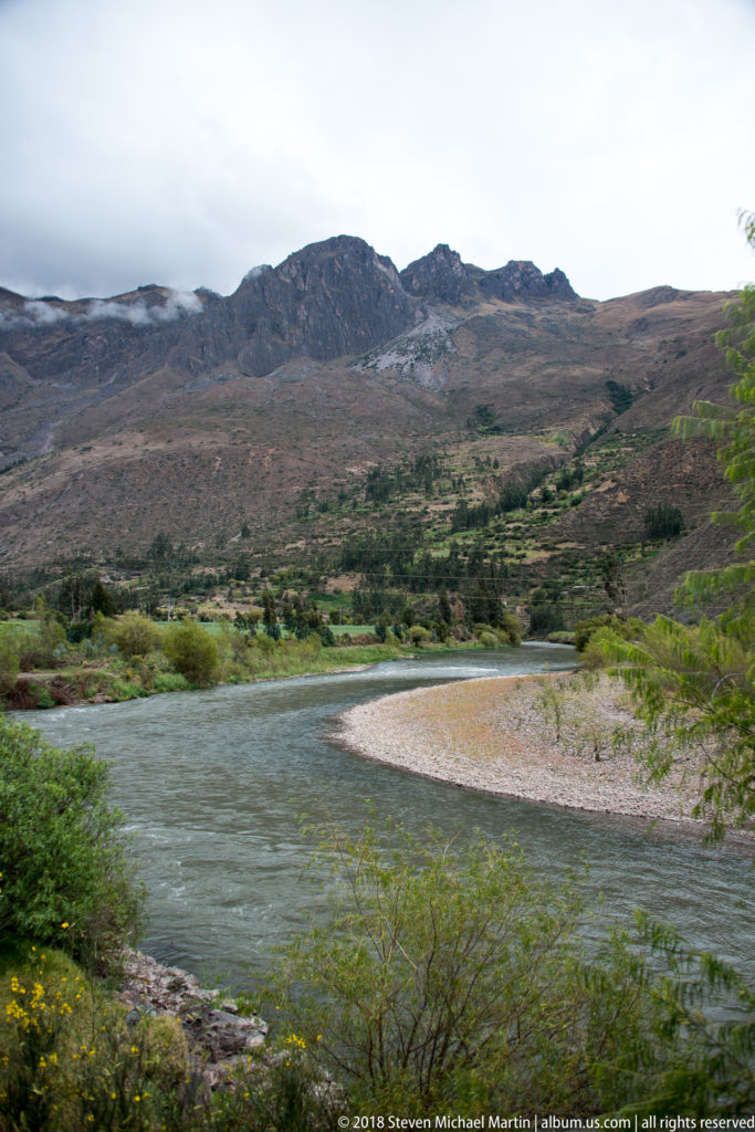 Greenish river curving around a pale gravel bar beneath broad, sparsely vegetated Andean ridges.