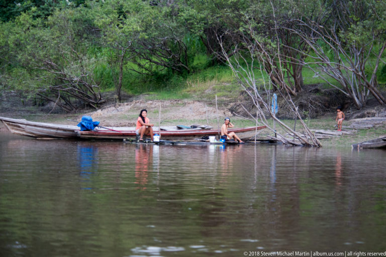 Several people beside long wooden canoes on a muddy riverbank, with buckets and household activity along the water.