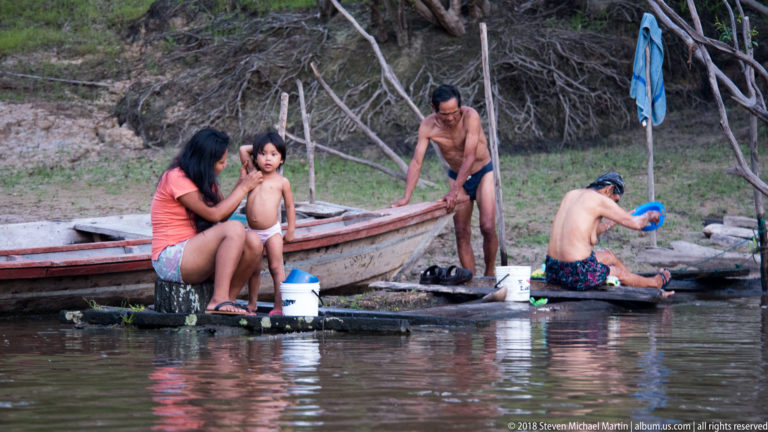 Adults and a child washing beside a long wooden canoe at the muddy riverbank, using buckets and containers.