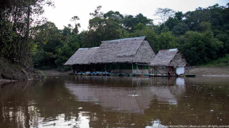 Long palm-thatched wooden building with smaller annex and narrow boats positioned directly along a muddy forest river.